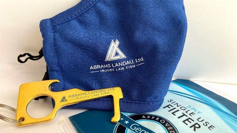 Blue face mask with ABRAMS LANDAU, Ltd., INJURY LAW FIRM stamped on it, mask filter packet, and contactless metal door opener. The VWCC's masking requirement has been updated.